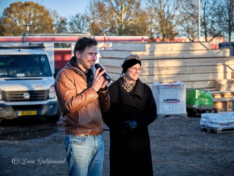 Twee mensen waarvan een met microfoon staan bij bouwmaterialen en lachen.