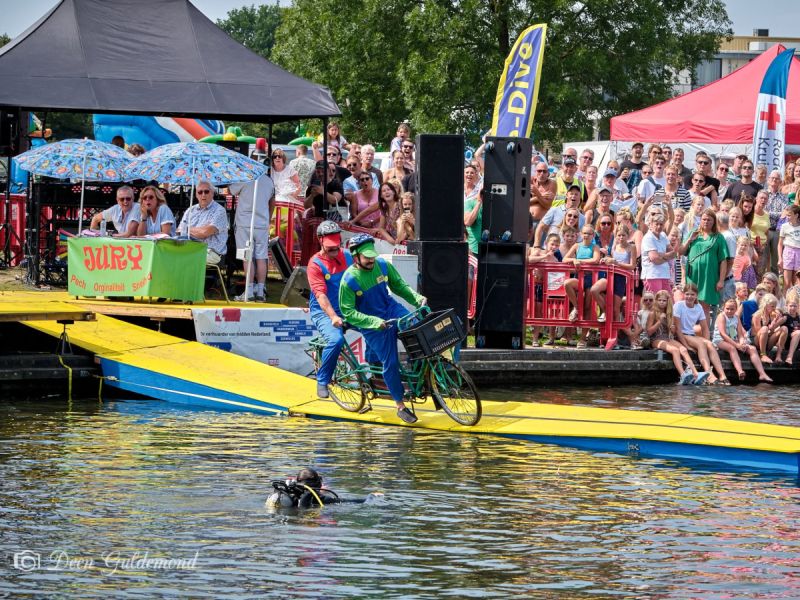 Mario en Luigi op de fiets die overhellen om in het water te vallen vanaf de baan. De jury en veel publiek aan de waterkant kijkt toe