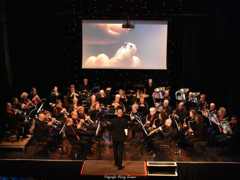 De FlevoFanfare in The Lux tijdens het Proms Concert, de diregent staat er voor en op het scherm een scene van een film