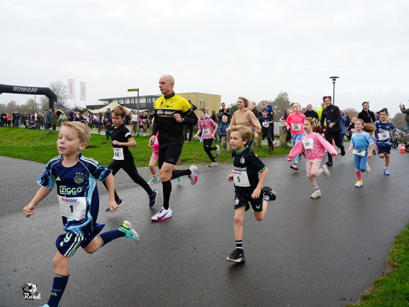 Jonge kinderen en ouderen die op de dijk de hardloopwedstrijd lopen met de brasserie op de achtergrond