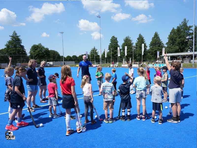 Een groep kleine kinderen voor een hockeytrainer op het blauwe veld van de hockey