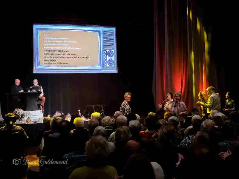 De ouderen kijkend naar het scherm op het podium voor de tekst waar ook Henk van Benthem en Han Koopman staan