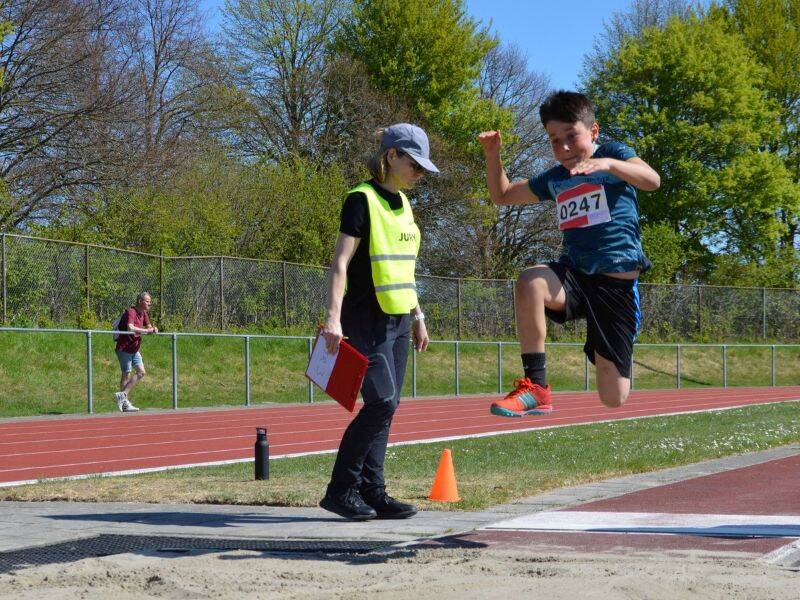 Kind met sportkleding springt in de lucht iemand in geel hesje met clipboard staat er naast