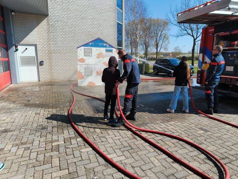mensen met een brandweerslang naast brandweerauto die de waterstraal richten op een huisje