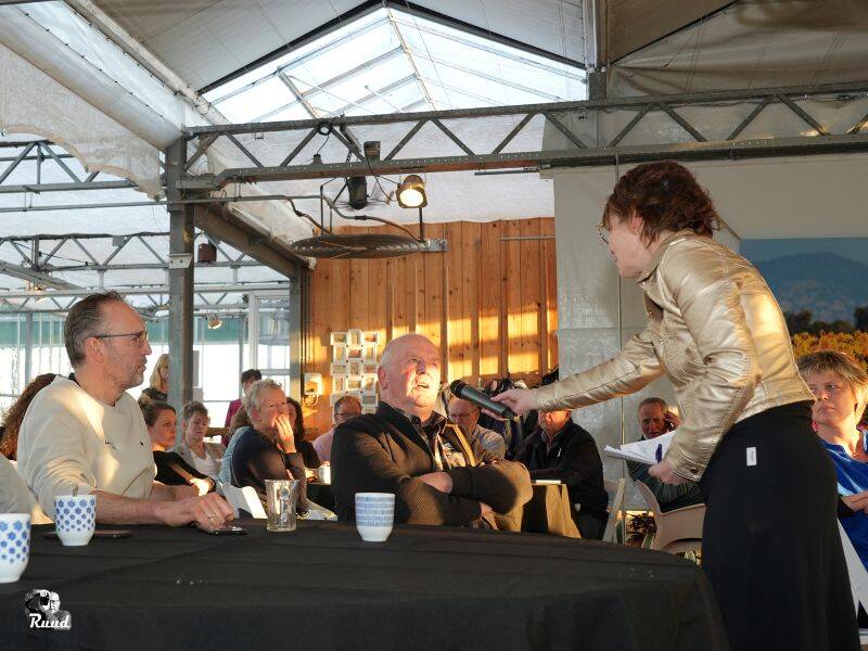 Verschillende mensen zitten aan een tafel met zwart kleed een praat in een microfoon die hem voorgehouden wordt