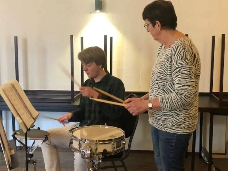 Het is vrijdag avond tegen 8 uur. De Kogge stroomt weer vol. Er klinken toonladders door het geroezemoes heen. Cindy zet er een lekkere beat onder middels het drumstel. Maar Catharine hoeft haar hand maar te heffen of het is stil. De derde bijeenkomst van het Nieuw Talenten Orkest is begonnen. En we pakken het steeds beter aan. Starten nu met een warming-up! De blazers blazen langzame tonen. De slagwerkers wordt aanbevolen een beetje te stretchen met de armen.  We repeteren de voorgaande oefeningen. Eerst in een langzaam tempo en dan steeds sneller. Catharina bespreekt de lipspanning, waardoor je verschillende tonen kunt maken. Iets waar de slagwerkgroep, de keyboard-spelers en violisten niks mee hoeft uiteraard. Maar des te belangrijker is die lipspanning voor alle saxofonisten, klarinettisten, hoornisten en trompettisten. We pakken een volgende oefening. Catharine is tevreden: “ Volgens mij hebben jullie heel goed geoefend.” Het klinkt als muziek in de oren.   Heel goede methode  Corien Donken is een collega-slagwerker. We staan naast elkaar op de snare-drum te trommelen. Corien zit in het onderwijs en organiseerde op haar school een tijdje geleden een trommeldag. Toen had ze de smaak te pakken. Ze is zeer enthousiast over de cursus: “ Het is een hele goede methode. Knap hoe Guus Pieksma dat doet. “ )Guus is de ‘uitvinder ‘ van het Nieuw Talenten Orkest.) Ze moet er echt even voor gaan zitten, voor het oefenen thuis. Maar de uitlegfilmpjes inspireren: “ Daar ben ik gek op.” Het is  niet Corien ’s eerste kennismaking met muziek. Ze speelde blokfluit, orgel en dwarsfluit. Maar ze wilde eens wat anders. “ Je kunt het overal doen,” aldus Corien. En dat zou ze ook best willen. “ Oh,” vraag ik ietwat verbaasd,” in een uniform marcherend?”  Corien lacht: “ Neeee… in een dweilorkest!” 