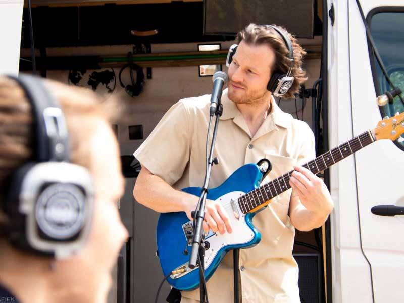 Guido vd Spek met een gitaar in de hand voor een bus en met koptelefoon op