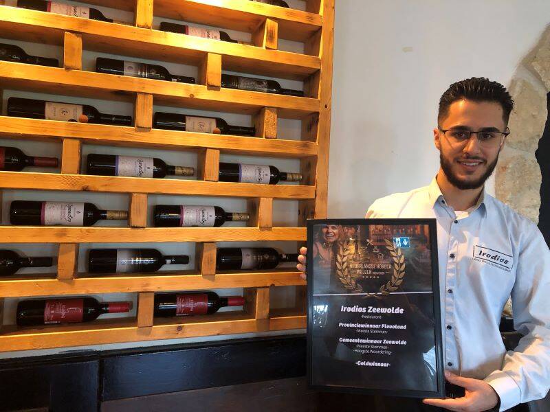 Hozhiin met de oorkonde in zijn hand bij het wijnrek van hout waarin flessen liggen