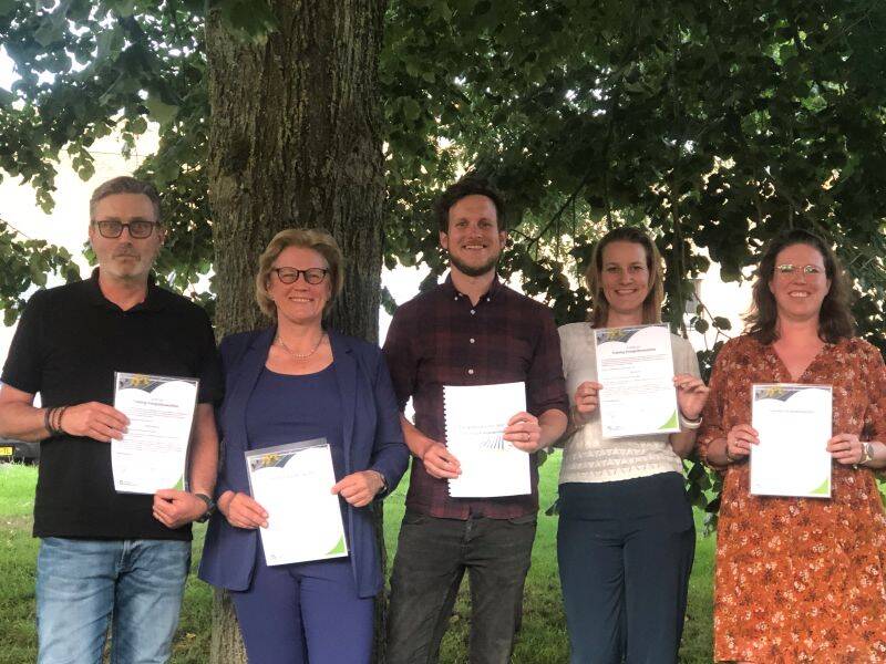 Vier mensen staan met een papier in hun handen onder een boom in een grasveld