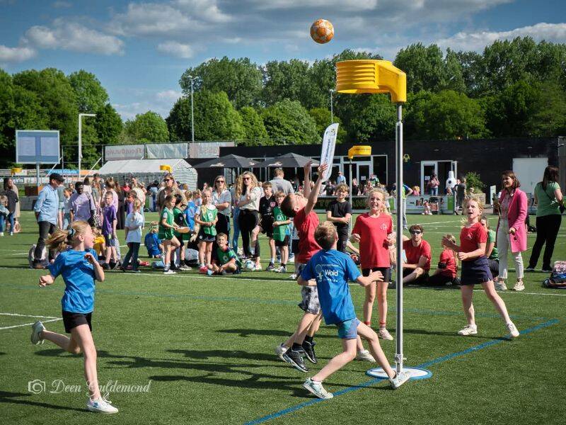 kinderen die korfballen op een grasveld met bomen en blauwe lucht op achtergrond