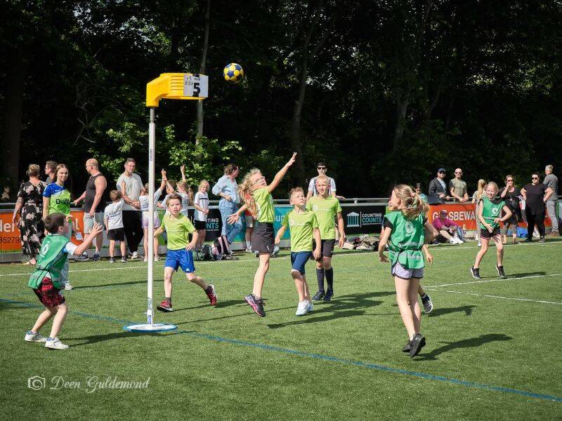 kinderen die korfballen op een grasveld met bomen en blauwe lucht op achtergrond