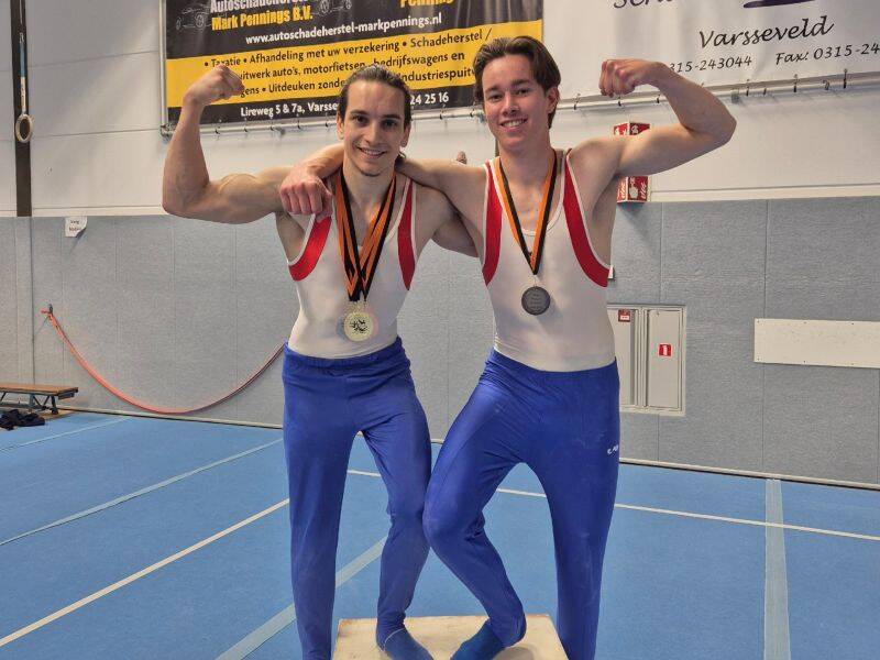 twee turners in blauwe broek met wit shirt staan op een houten blok met medailles om hun nek